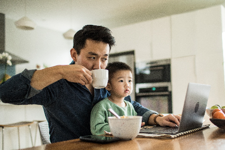 A father takes a sip of coffee while seated at the table with his young son in his lap and his laptop open in front of him.